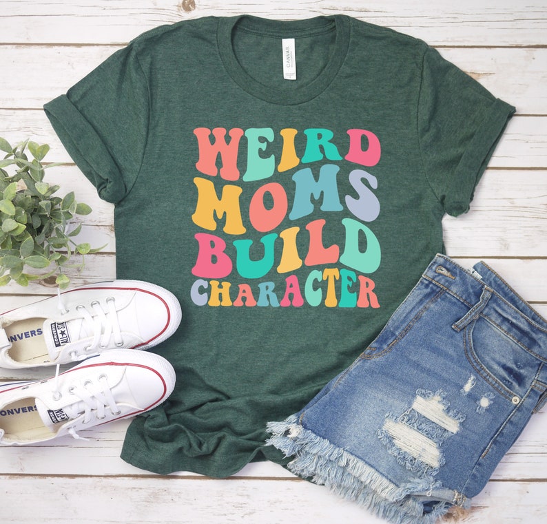 Pode incluir: Uma camiseta verde com o texto "Weird Moms Build Character" em fonte retr&ocirc; colorida.