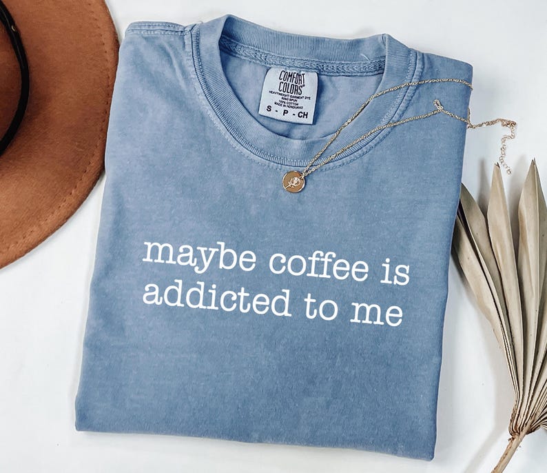 May include: A blue Comfort Colors t-shirt with the phrase "maybe coffee is addicted to me" in white lettering. A gold necklace with a pendant is draped across the shirt. A brown hat and dried plant are in the background.