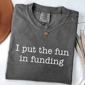 May include: A dark gray t-shirt with the phrase "I put the fun in funding" printed in white. A gold-colored necklace with a pendant rests on the shirt. A brown hat and dried plant are in the background.
