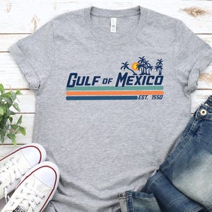 May include: A gray t-shirt with a retro-style graphic that says "Gulf of Mexico Est. 1950" with palm trees and a sunset. The shirt is paired with blue jeans and white sneakers.