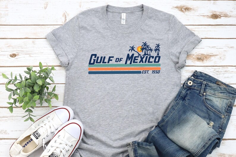 May include: A gray t-shirt with a retro-style graphic that says "Gulf of Mexico Est. 1950" with palm trees and a sunset. The shirt is paired with blue jeans and white sneakers.
