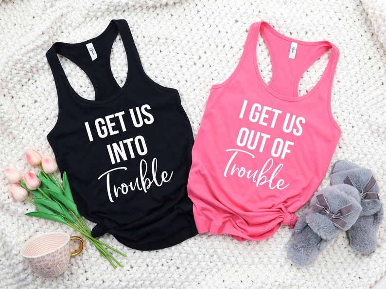 Pode incluir: Duas blusas de al&ccedil;as pretas com texto branco. A blusa &agrave; esquerda diz "I get us into trouble". A blusa &agrave; direita diz "I get us out of trouble".