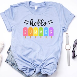 May include: A light blue t-shirt with the text "hello summer" in black script.  The text is surrounded by six rainbow popsicle graphics.