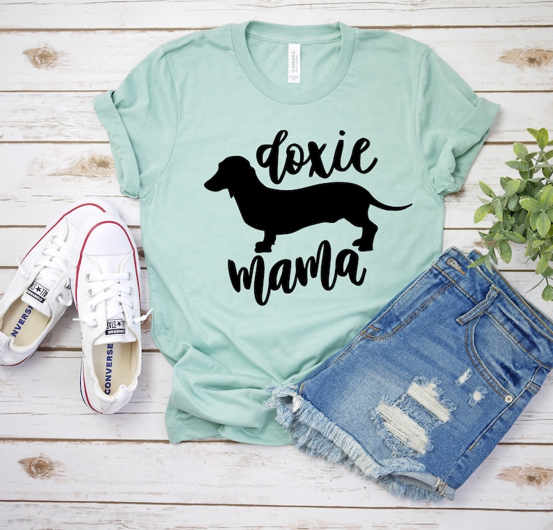 May include: A light blue t-shirt with a black silhouette of a dachshund and the text "doxie mama". The shirt is laying on a white wooden surface with a pair of white sneakers and a pair of blue denim shorts.