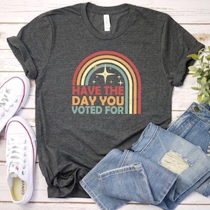 May include: Dark gray t-shirt with a retro rainbow design and the text "Have the Day You Voted For." The shirt is paired with blue jeans and white canvas sneakers.
