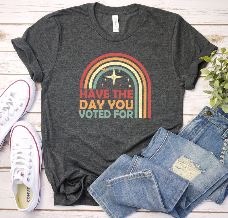 May include: Dark gray t-shirt with a retro rainbow design and the text "Have the Day You Voted For." The shirt is paired with blue jeans and white canvas sneakers.