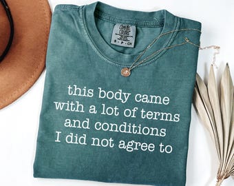 Op de afbeelding: Een blauwgroen T-shirt met korte mouwen en de witte tekst: this body came with a lot of terms and conditions I did not agree to. Het shirt heeft een Comfort Colors merklabel bij de binnenhals en een kleine goudkleurige ketting.