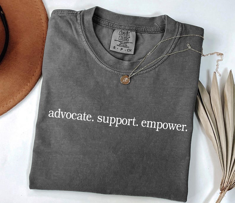 Op de afbeelding: Een donkergrijs t-shirt met de woorden "advocate. support. empower." in het wit. Een zilveren ketting met een hanger ligt op het shirt. Een bruine hoed en gedroogde bladeren zijn op de achtergrond.