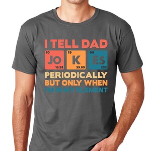 Puede incluir: Camiseta gris oscuro con la frase "I TELL DAD JOKES PERIODICALLY BUT ONLY WHEN I'M IN MY ELEMENT" en un dise&ntilde;o de tabla peri&oacute;dica. La camiseta tiene cuello redondo y mangas cortas.