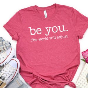 Puede incluir: Una camiseta rosa con la frase "be you. The world will adjust" en blanco. Se combina con vaqueros azules y zapatillas blancas. Una peque&ntilde;a bolsa de regalo y rosas rosas completan el conjunto.