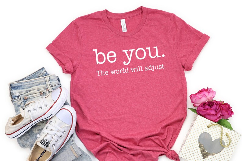 Puede incluir: Una camiseta rosa con la frase "be you. The world will adjust" en blanco. Se combina con vaqueros azules y zapatillas blancas. Una peque&ntilde;a bolsa de regalo y rosas rosas completan el conjunto.