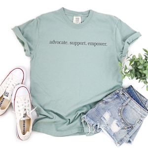 Op de afbeelding: Een lichtgroen t-shirt met de woorden "advocate. support. empower." in zwarte letters. Het shirt wordt gecombineerd met een spijkerbroek, witte sneakers en een kleine groene plant.