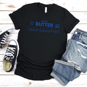 May include: Black t-shirt with a blue graphic that says "SALTED BUTTER GRADE AA" and a chart with measurements. The shirt is laid flat on a white surface with a pair of black sneakers and blue jeans.