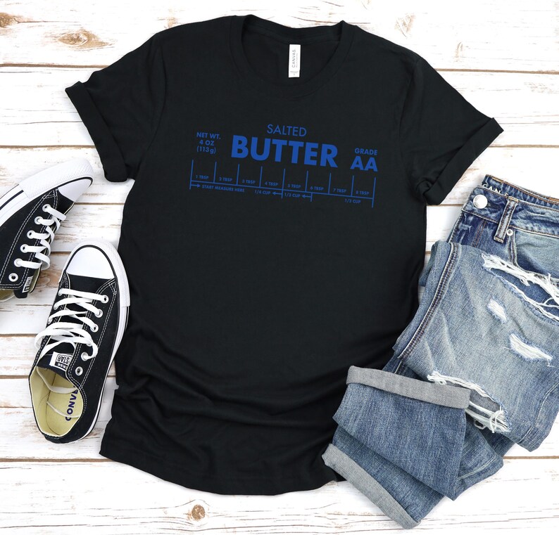 May include: Black t-shirt with a blue graphic that says "SALTED BUTTER GRADE AA" and a chart with measurements. The shirt is laid flat on a white surface with a pair of black sneakers and blue jeans.