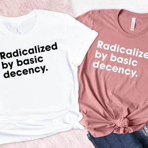 May include: Two t-shirts, one white and one dusty rose, with the phrase "Radicalized by basic decency" printed in black and white. The shirts are displayed on a pink surface with a pair of blue jeans.