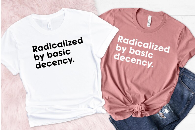 May include: Two t-shirts, one white and one dusty rose, with the phrase "Radicalized by basic decency" printed in black and white. The shirts are displayed on a pink surface with a pair of blue jeans.