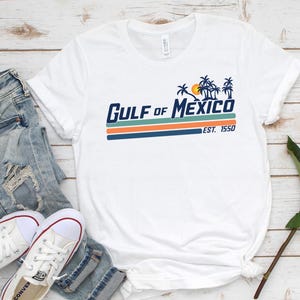 May include: White t-shirt with a blue and orange graphic that says "GULF of MEXICO EST. 1550". The graphic features palm trees and a sun.