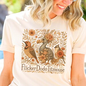 May include: A beige t-shirt with a graphic print of a dodo bird, a flicker bird, and a titmouse surrounded by flowers and leaves. The text "Flicker Dodo Titmouse" is printed below the graphic.