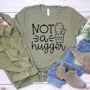 May include: A green t-shirt with the text "NOT a hugger" and a cactus graphic. The shirt is laid out on a white wooden surface with a green jacket, blue jeans, and brown boots.