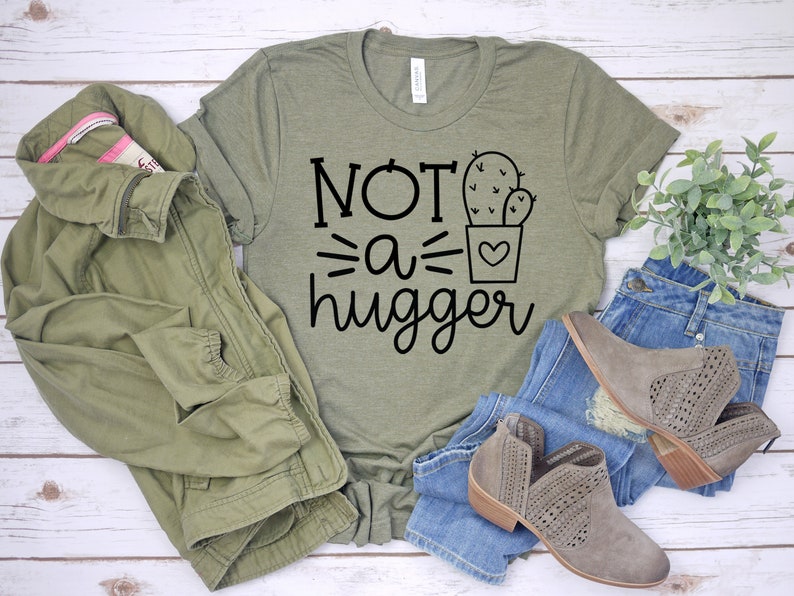 May include: A green t-shirt with the text "NOT a hugger" and a cactus graphic. The shirt is laid out on a white wooden surface with a green jacket, blue jeans, and brown boots.