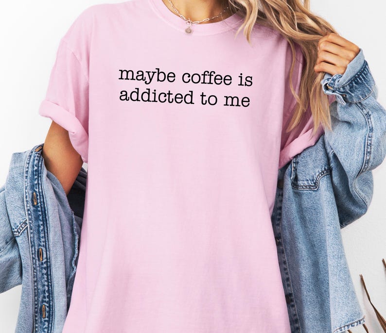 May include: A light pink t-shirt with the text "maybe coffee is addicted to me" in black. The shirt has short sleeves and a crew neck. A denim jacket is draped over the shoulders.