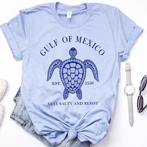 Puede incluir: Camiseta azul claro con un gr&aacute;fico azul marino de una tortuga marina y el texto "GULF OF MEXICO EST. 1550 STAY SALTY AND RESIST". La camiseta se combina con vaqueros, zapatos, un reloj y gafas de sol.