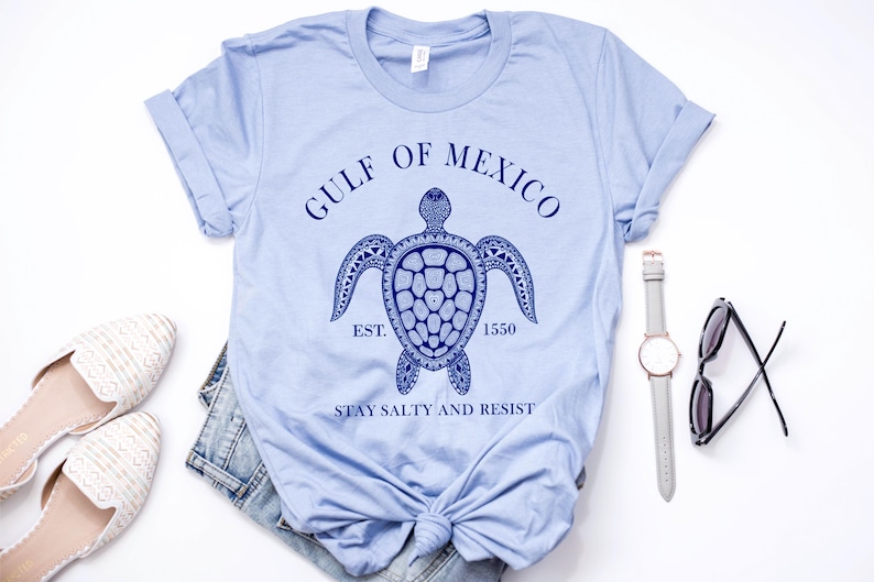 Puede incluir: Camiseta azul claro con un gr&aacute;fico azul marino de una tortuga marina y el texto "GULF OF MEXICO EST. 1550 STAY SALTY AND RESIST". La camiseta se combina con vaqueros, zapatos, un reloj y gafas de sol.