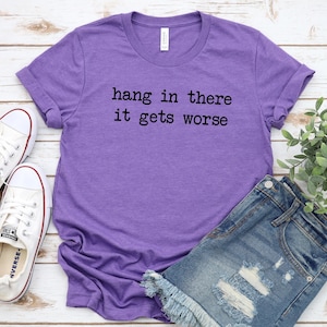 May include: A purple t-shirt with the text "hang in there it gets worse" in black lettering. The shirt is paired with denim shorts and white sneakers with red accents. A sprig of greenery is in the background.