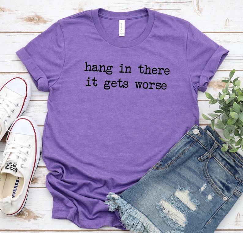 May include: A purple t-shirt with the text "hang in there it gets worse" in black lettering. The shirt is paired with denim shorts and white sneakers with red accents. A sprig of greenery is in the background.