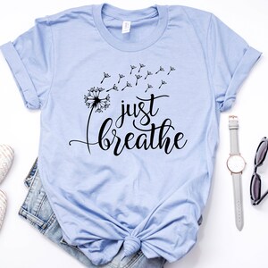 Op de afbeelding: Een lichtblauw t-shirt met een zwarte paardenbloem afbeelding en de tekst "just breathe" in een cursief schrift.