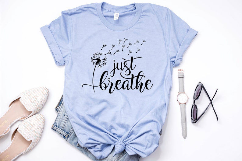 Op de afbeelding: Een lichtblauw t-shirt met een zwarte paardenbloem afbeelding en de tekst "just breathe" in een cursief schrift.