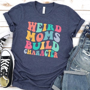 Pode incluir: Uma camiseta azul marinho com o texto "Weird Moms Build Character" em cores do arco-&iacute;ris. O texto est&aacute; em um estilo de fonte retr&ocirc;.