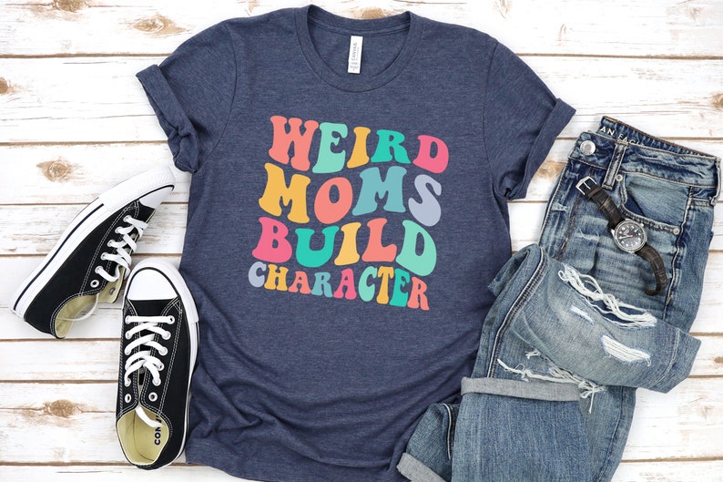 Pode incluir: Uma camiseta azul marinho com o texto "Weird Moms Build Character" em cores do arco-&iacute;ris. O texto est&aacute; em um estilo de fonte retr&ocirc;.