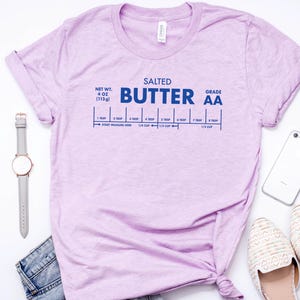 May include: A light purple t-shirt with a graphic of a stick of butter with the text "SALTED BUTTER GRADE AA" and a measurement guide for tablespoons and cups. The t-shirt is on a white surface with a watch, a phone, and a pair of white shoes.