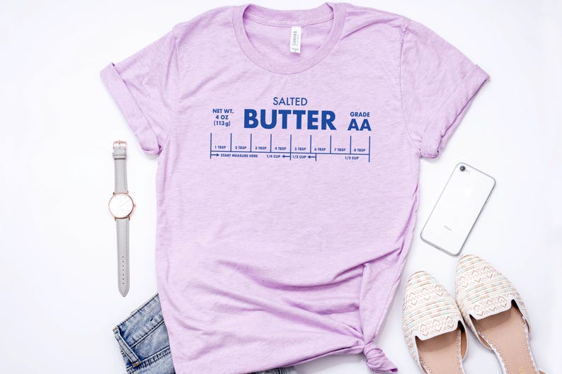 May include: A light purple t-shirt with a graphic of a stick of butter with the text "SALTED BUTTER GRADE AA" and a measurement guide for tablespoons and cups. The t-shirt is on a white surface with a watch, a phone, and a pair of white shoes.