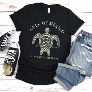 Puede incluir: Camiseta negra con un gr&aacute;fico de tortuga crema y el texto "GULF OF MEXICO EST. 1550 STAY SALTY AND RESIST." La imagen incluye vaqueros azules y zapatillas deportivas blancas y negras.