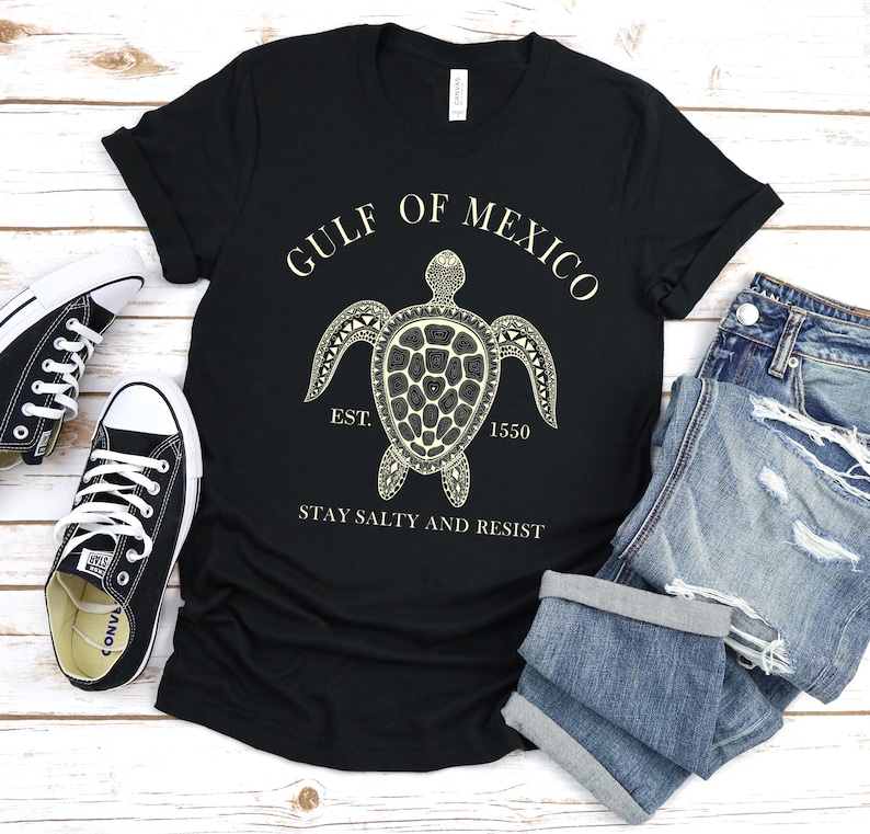 Puede incluir: Camiseta negra con un gr&aacute;fico de tortuga crema y el texto "GULF OF MEXICO EST. 1550 STAY SALTY AND RESIST." La imagen incluye vaqueros azules y zapatillas deportivas blancas y negras.