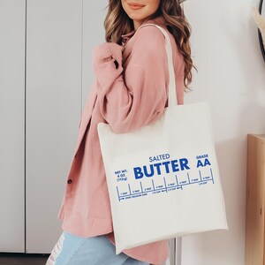 May include: A white canvas tote bag with a blue graphic that reads "Salted Butter Grade AA" and includes a measurement guide for tablespoons and cups. The bag is being held by a person wearing a pink sweater.