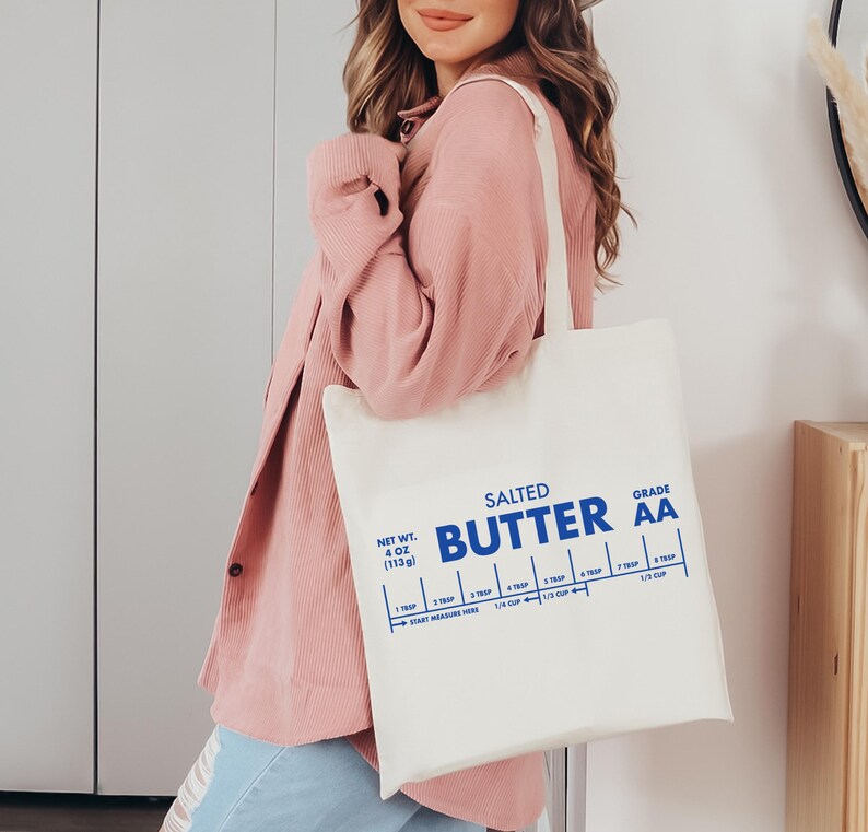 May include: A white canvas tote bag with a blue graphic that reads "Salted Butter Grade AA" and includes a measurement guide for tablespoons and cups. The bag is being held by a person wearing a pink sweater.