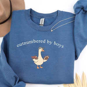 May include: A blue sweatshirt with the text "outnumbered by boys" and a cartoon goose wearing a baseball cap, holding a football and a toy truck. A gold necklace with a star pendant is also visible.