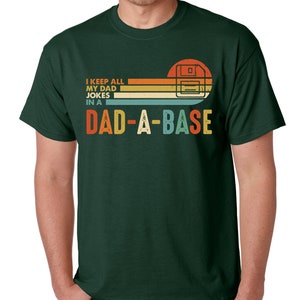 Op de afbeelding: Donkergroen t-shirt met een retro design. Het ontwerp toont een floppy disk-afbeelding en de tekst "I KEEP ALL MY DAD JOKES IN A DAD-A-BASE" in oranje, geel en blauw. Het shirt heeft een klassieke ronde hals.