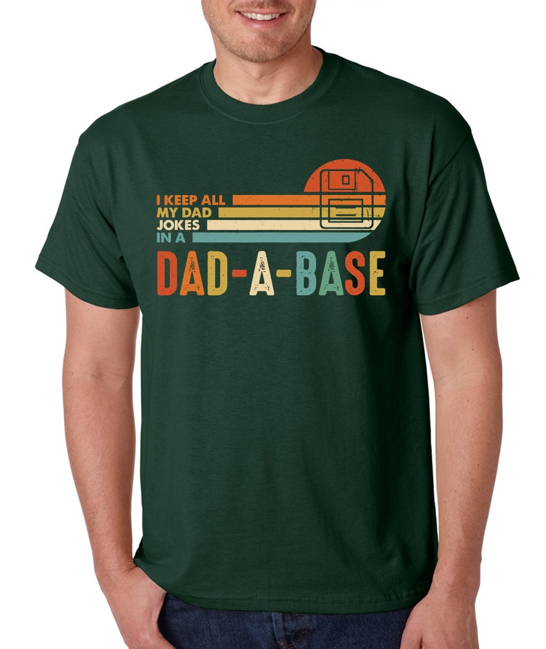 Op de afbeelding: Donkergroen t-shirt met een retro design. Het ontwerp toont een floppy disk-afbeelding en de tekst "I KEEP ALL MY DAD JOKES IN A DAD-A-BASE" in oranje, geel en blauw. Het shirt heeft een klassieke ronde hals.