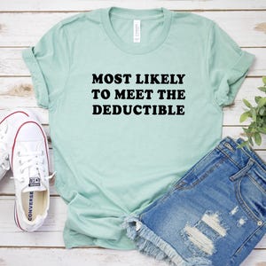 May include: A light green t-shirt with the text "MOST LIKELY TO MEET THE DEDUCTIBLE" in black. The shirt is paired with denim shorts and white sneakers. The shirt is made of a soft material and has rolled-up sleeves.