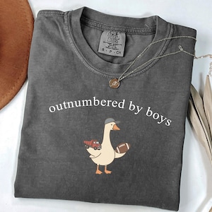 May include: Dark grey Comfort Colors t-shirt with the text "outnumbered by boys". A cartoon goose wearing a cap and holding a football and a truck is printed on the front. A silver necklace with a pendant is on the shirt.