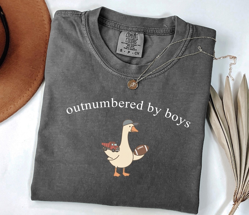 May include: Dark grey Comfort Colors t-shirt with the text "outnumbered by boys". A cartoon goose wearing a cap and holding a football and a truck is printed on the front. A silver necklace with a pendant is on the shirt.