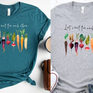 May include: Two t-shirts with a watercolor design of vegetables and the text "Let's root for each other". The shirts are teal and gray.
