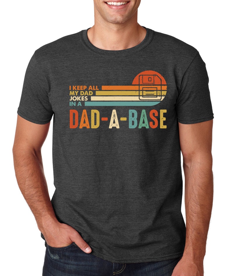 Op de afbeelding: Donkergrijs t-shirt met een retro design. Het shirt heeft de tekst "I KEEP ALL MY DAD JOKES IN A DAD-A-BASE" in een kleurrijk, vintage lettertype. Het ontwerp bevat een gestileerde afbeelding van een diskette.