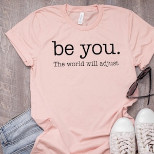 Puede incluir: Una camiseta rosa claro con las palabras "be you." y "The world will adjust" impresas en negro. La camiseta se combina con vaqueros azules, gafas de sol y zapatillas blancas con detalles grises. Los art&iacute;culos est&aacute;n colocados sobre una superficie de madera.