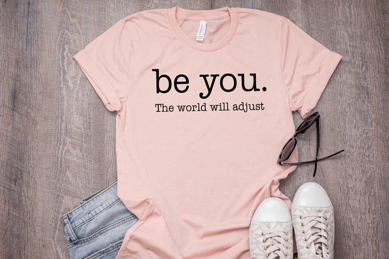 Puede incluir: Una camiseta rosa claro con las palabras "be you." y "The world will adjust" impresas en negro. La camiseta se combina con vaqueros azules, gafas de sol y zapatillas blancas con detalles grises. Los art&iacute;culos est&aacute;n colocados sobre una superficie de madera.