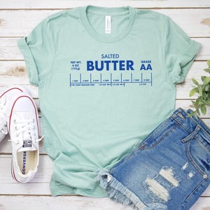 May include: A light blue t-shirt with the text "SALTED BUTTER AA" in blue lettering. The shirt also includes a graphic of a butter stick with measurements in tablespoons and cups.